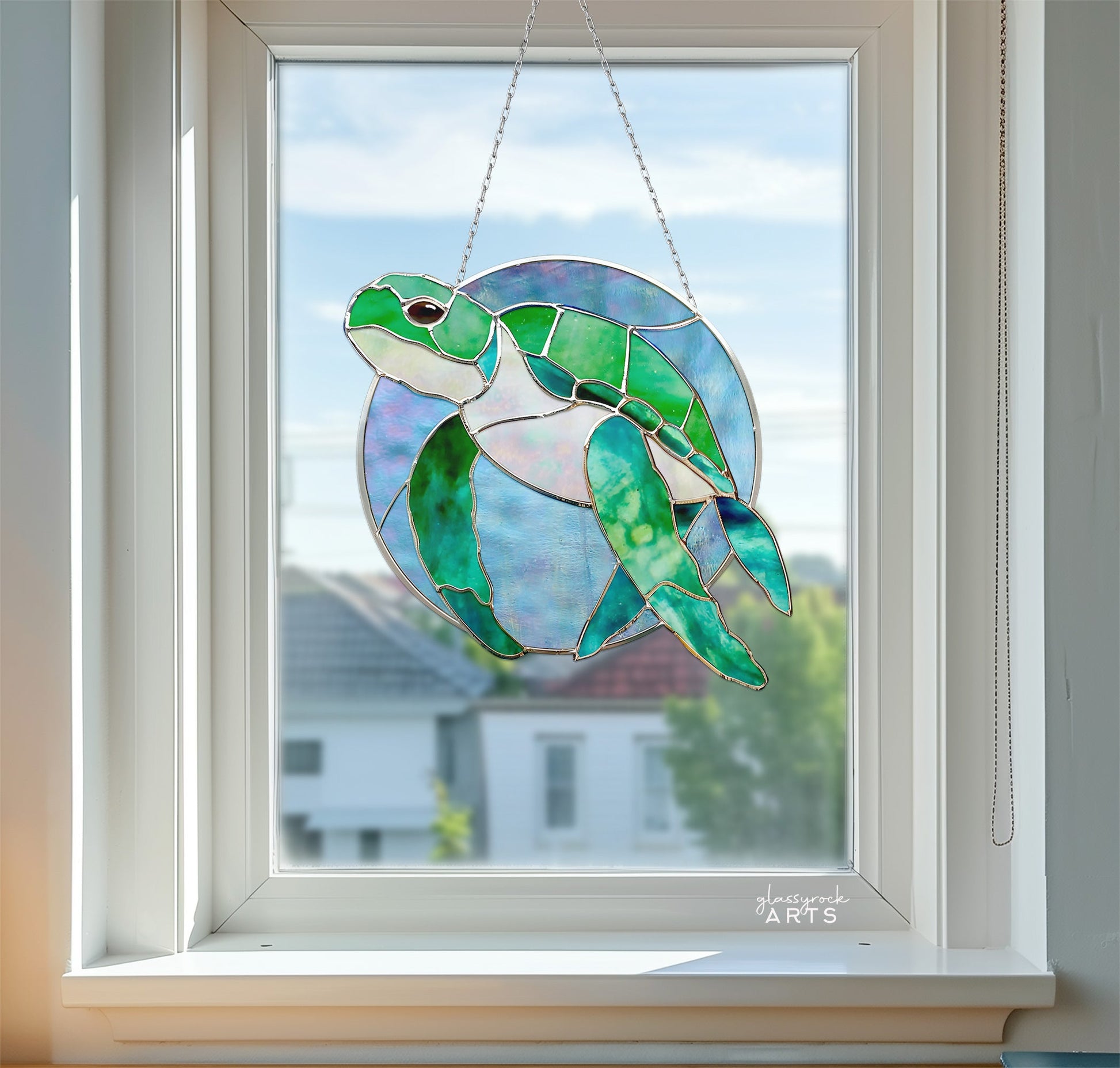 The Round Stained Glass Turtle Pattern, in green and blue, graces the window. Sunlight illuminates its colors while a soft blur of houses and trees outside adds charm to this elegant decor.