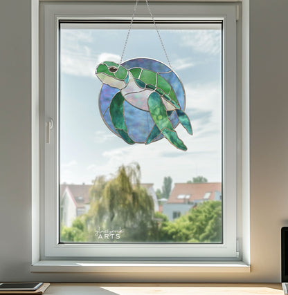 In a window, the Round Stained Glass Turtle Pattern sun catcher hangs elegantly, showcasing a vibrant green sea turtle against a blue circular background, with blurred trees and houses visible against a clear sky.