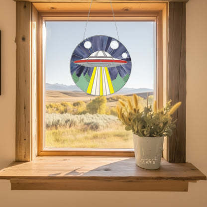 A wooden-framed window reveals fields and distant mountains under a blue sky. A UFO Stained Glass Pattern sun catcher hangs there, casting colorful reflections, while a potted plant with yellow flowers on the sill adds a natural touch to the peaceful scene.