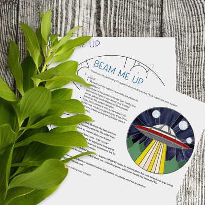 A UFO Stained Glass Pattern sun catcher sits on a gray wooden surface next to an instruction sheet titled BEAM ME UP, with lush green leaves enhancing the modern design.