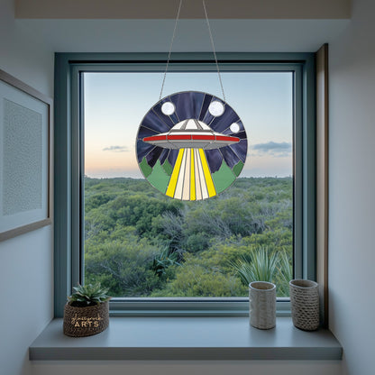 The UFO Stained Glass Pattern sun catcher shines vibrantly in a window, casting colorful beams over a lush green forest beneath a cloudy sky. Two small potted plants on the sill add charm and complement its whimsical design.