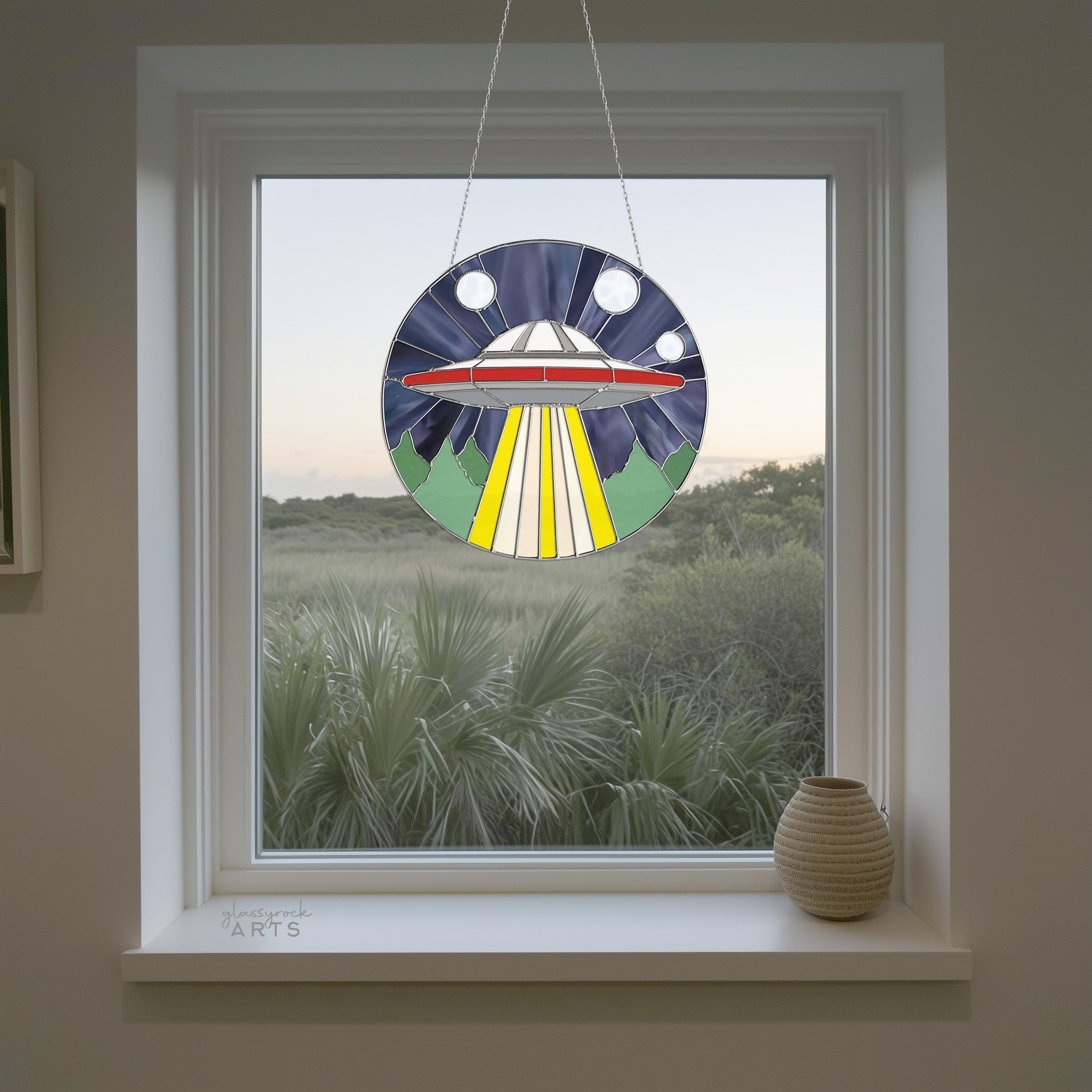 The UFO Stained Glass Pattern features a vibrant sun catcher that gracefully hangs in a window, with a green landscape of bushes and trees outside and a small vase on the sill, offering a stunning modern design sure to captivate any observer.