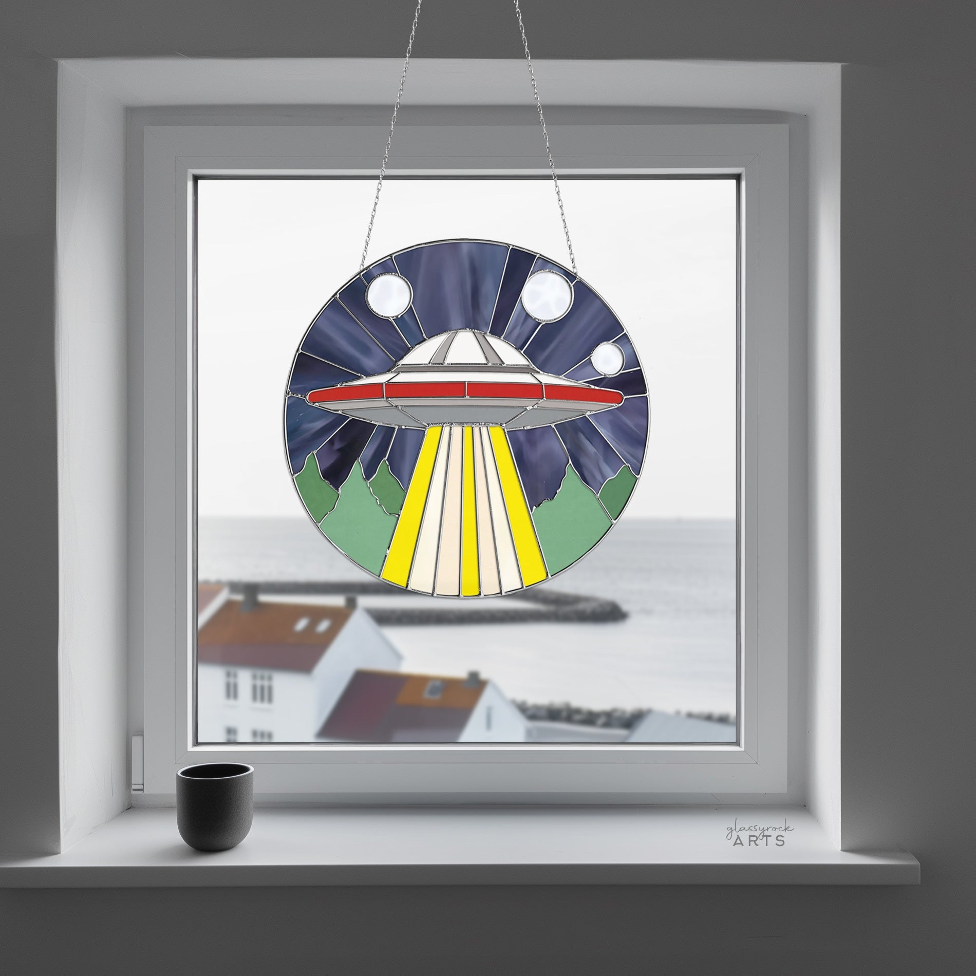 The UFO Stained Glass Pattern hangs as a sun catcher in a window with a coastal view of white houses and sea, complemented by a black mug on the windowsill.