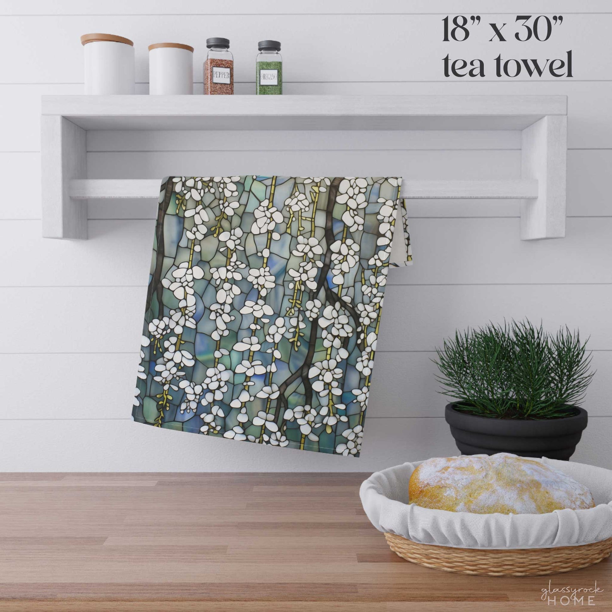 A Stained Glass White Wisteria Dish Towel, featuring a multicolored floral pattern and crafted from easy-care polyester, hangs on a white shelf that also holds two jars and two white containers. Below, a green plant and a bowl with cloth rest on a wooden surface.