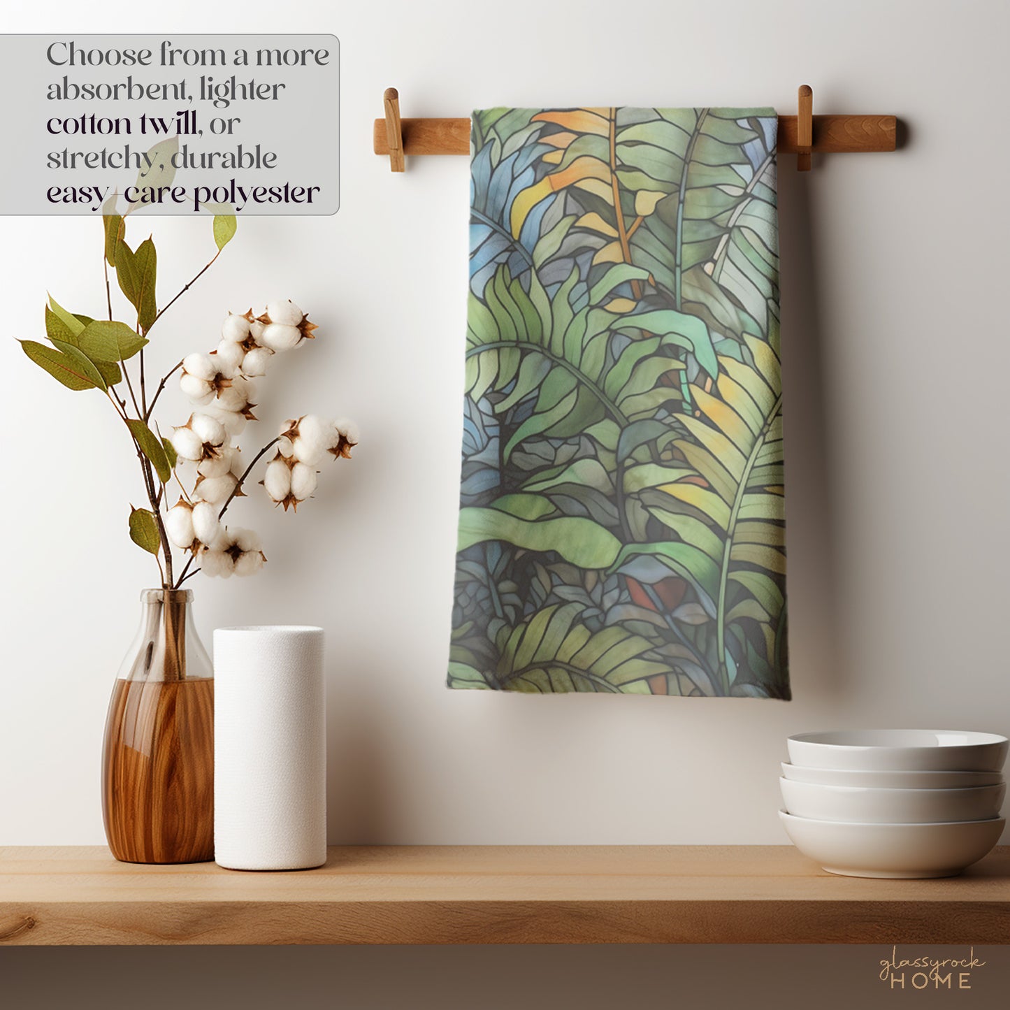 A kitchen scene features the Stained Glass Wild Ferns Dish Towel hanging on a wooden peg. Nearby, a vase with cotton stems, a paper towel roll, and stacked white bowls complete the look. Choose from absorbent cotton twill or easy-care polyester for your custom creation.