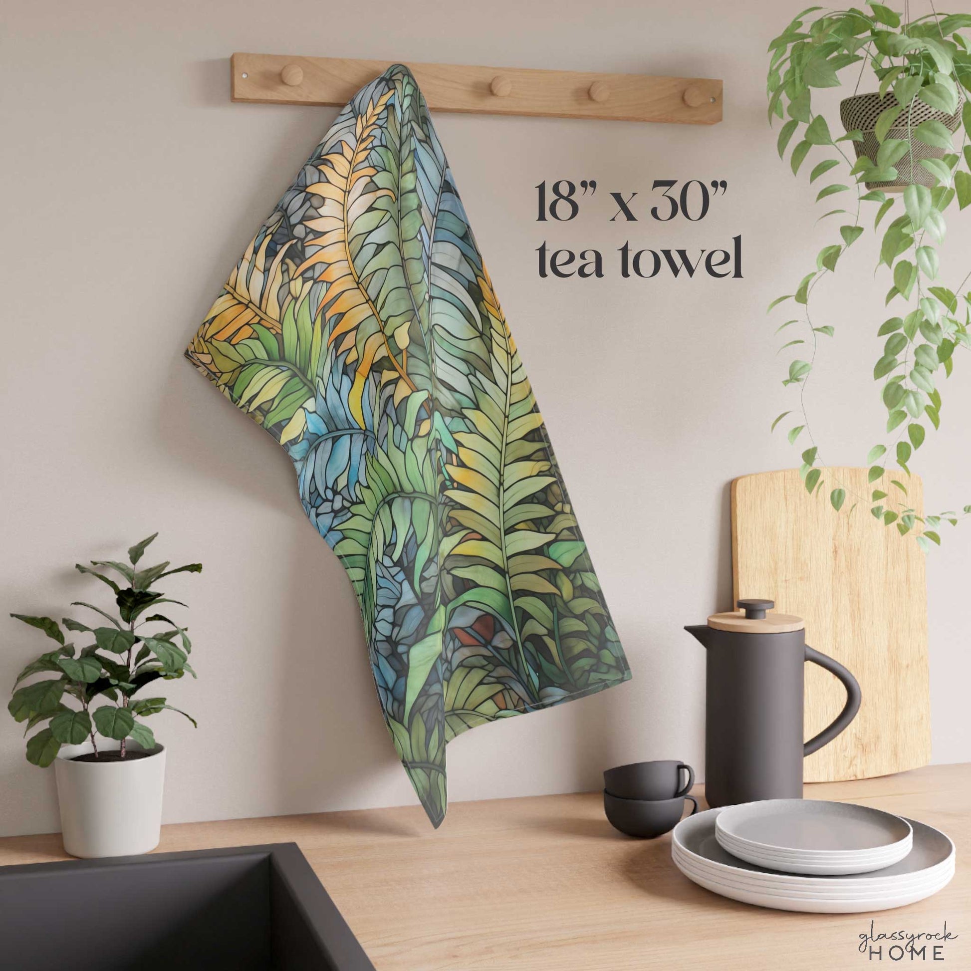 An 18 x 30 Stained Glass Wild Ferns Dish Towel hangs on a wooden rack. Nearby, a potted plant, black kettle, cup, and stacked plates sit on the light wood countertop.