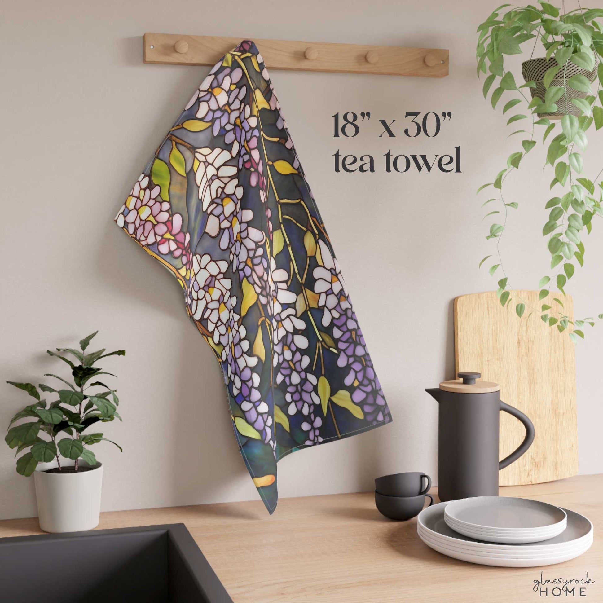 The Stained Glass Wisteria Dish Towel, a vibrant natural cotton twill tea towel featuring a white wisteria design, hangs on a wooden peg in a modern kitchen. Text on the wall reads "18” x 30” tea towel." Nearby are a potted plant, a black teapot, cups, and plates.