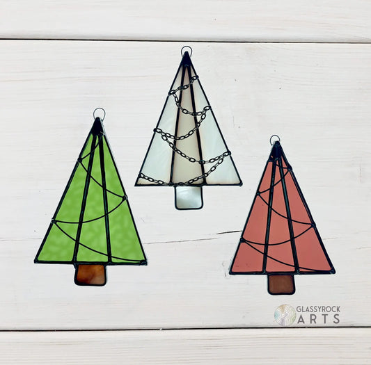 Three Handmade Christmas Tree Suncatchers in green, white, and red with black outlines and decorative patterns are displayed against a light wooden background, showcasing Noelle Barnes artistry. The Glassyrock Arts logo appears at the bottom right.