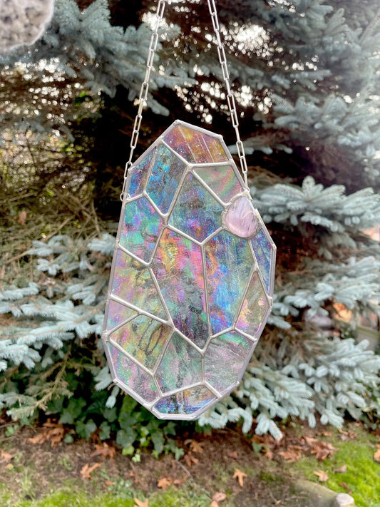 The Free Giant Gem, a stained glass suncatcher with an abstract geometric design by Noelle Barnes of GlassyRock Arts, hangs from a chain. It displays colorful iridescent hues against green pine tree branches. Use the code from your email at checkout.